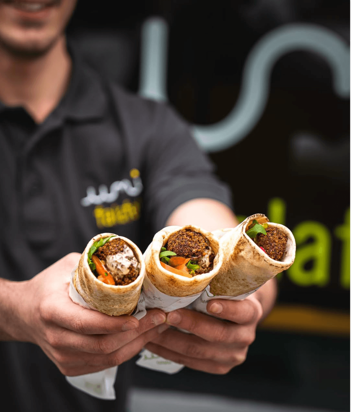 Three falafel wraps held out in front of an Umi Falafel shopfront